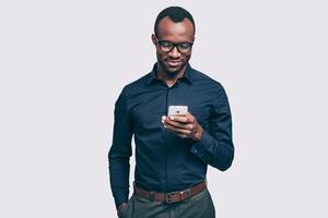 Typing quick message. Handsome young African man holding smart phone and looking at it while standing against grey background photo
