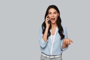 Good business talk. Surprised young woman talking on the phone while standing against grey background photo