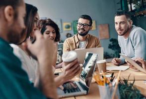 Collaboration is a key to best results. Group of young modern people in smart casual wear discussing something and smiling while working in the creative office photo