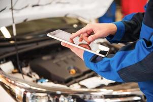 His helper at work. Close-up of man in uniform working on digital tablet while standing in front of car hood in workshop photo