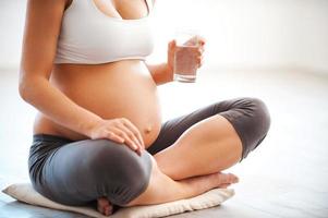 Maintaining water balance. Close-up of pregnant young woman holding glass of water while sitting in lotus position photo