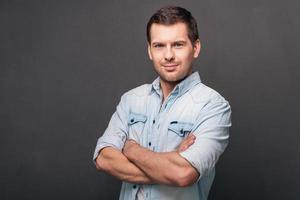 Mr. Confidence. Cheerful young man keeping arms crossed and looking at camera while standing against grey background photo