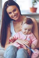 la mejor mamá del mundo. bella joven sosteniendo a una niña con un juguete de rodillas y mirando a la cámara con una sonrisa mientras se sienta en el sofá de casa foto