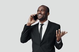 Happy young African man in formalwear talking on the smart phone and smiling while standing against grey background photo