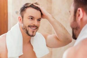 Good morning to me. Handsome young man touching his hair with hand and smiling while standing in front of the mirror photo
