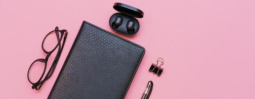 Black notebook with glasses and a pen, wireless headphones on a pink background. photo