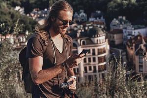 chatear con amigos, chateando con amigos. joven con ropa informal usando un teléfono inteligente y sonriendo mientras está de pie en la colina al aire libre foto