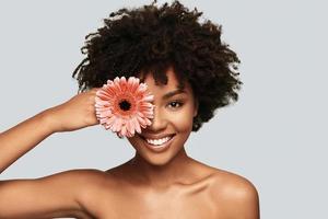 Dangerously beautiful. Attractive young African woman looking at camera and covering eye with a flower while standing against grey background photo
