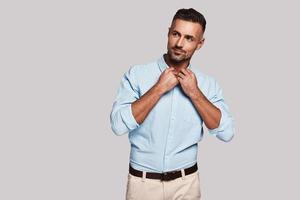 Elegant man. Good looking young man adjusting collar and smiling while standing against grey background photo