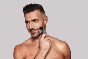 Enjoying morning routine. Charming young man shaving and smiling while standing against grey background photo