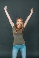 She is a winner Happy young woman with blond hair in casual wear keeping arms raised and looking at camera while standing against grey background photo