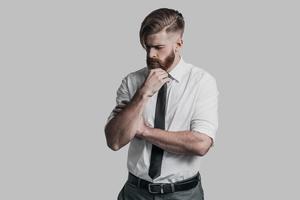 Time to make important decision. Thoughtful young businessman keeping hand on chin while standing against grey background photo