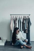 Thinking about the right choice. Thoughtful young woman in casual wear sitting on the floor at home and holding hand in hair near her clothes hanging on the racks photo