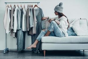 Maybe this one  Thoughtful young woman in casual wear choosing the dress while sitting on the couch at home near her clothes hanging on the racks photo