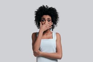 Surprised young African woman in casual wear covering mouth with hand and looking at camera while standing against grey background photo