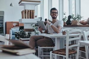 alegre y guapo. un joven apuesto y moderno que sostiene una tableta digital y mira hacia otro lado mientras se sienta en la mesa del café foto