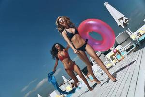 momentos de felicidad. Longitud total de dos hermosas mujeres jóvenes en bikini corriendo juntas cerca de la piscina mientras una de ellas lleva un anillo inflable foto