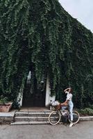 viviendo la vida verde. longitud completa de una joven atractiva con ropa informal manteniendo la mano en el cabello mientras está de pie frente a la casa cubierta de hiedra foto