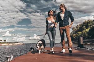 Love is in the air. Full length of beautiful young couple looking at each other and smiling while walking with their dog the river shore photo