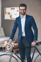 Modern businessman. Handsome young man wearing glasses and suit looking at camera with smile while standing with retro bicycle in office photo