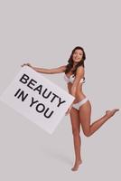 You were born beautiful.  Full length of attractive young woman holding a poster and looking at camera with smile while standing against grey background photo