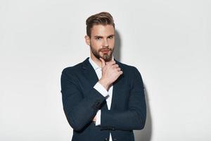 Used to look perfect. Handsome young man in full suit keeping hand on chin and looking at camera while standing against grey background photo