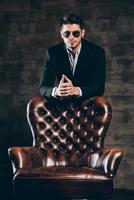 Charming and elegant. Young handsome man in suit and sunglasses leaning on leather chair and looking at camera while standing against dark grey background photo