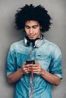 Searching for new song. Young African man wearing headphones and using his smartphone while standing against grey background photo