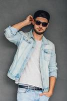 Modern and cool. Confident young man in sunglasses adjusting his baseball cap and looking at camera while standing against grey background photo