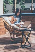 disfrutando de un buen rato al aire libre. bella joven trabajando en su laptop y sonriendo mientras se sienta en una silla grande y cómoda en la terraza de su casa al aire libre foto