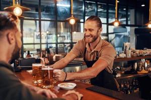 encantador joven barman en delantal sirviendo cerveza y sonriendo mientras trabaja en el pub foto