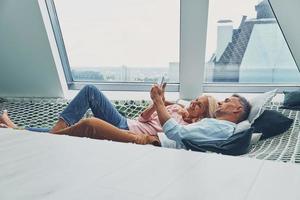 Beautiful mature couple examining smart phone and smiling while relaxing in big hammock at home together photo