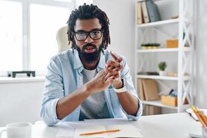 Confident young African man in eyewear telling something and looking at camera while working indoors photo