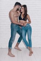 Full length of beautiful young couple looking at each other and smiling while standing against brick wall photo