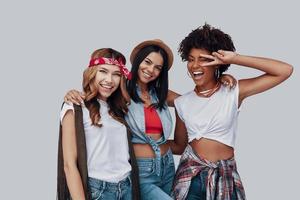 tres atractivas mujeres jóvenes con estilo mirando a la cámara y sonriendo mientras están de pie contra el fondo gris foto