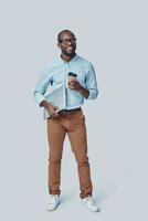 Full length of confident young African man carrying laptop and smiling while standing against grey background photo