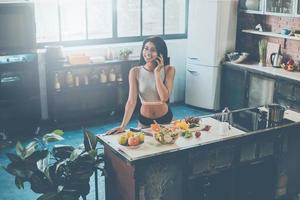 charla despreocupada con un amigo. vista superior de una joven de raza mixta hablando por teléfono móvil y sonriendo mientras prepara la comida en la cocina foto