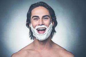 Shaving is fun. Portrait of young shirtless man with shaving cream on face looking at camera and smiling while standing against grey background photo