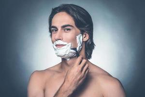 Morning shave. Handsome young shirtless man shaving and looking at camera while standing against grey background photo