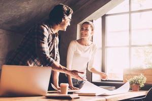 Working on successful project together. Cheerful young man and woman in casual wear pointing blueprint and communicating while both standing near wooden desk in creative office photo