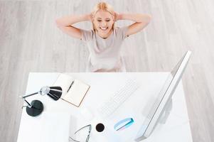 Taking a minute break. Top view of beautiful young smiling woman holding hands behind head while sitting at her working place photo