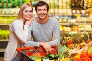 ir de compras juntos para la cena. alegre pareja joven sonriendo a la cámara y de pie detrás de su carrito de compras en una tienda de alimentos foto