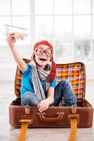 Dreaming of a sky. Happy little boy in pilot headwear and eyeglasses playing with wooden planer and smiling while sitting inside of briefcase at home photo