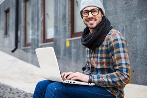 tener oportunidad de trabajar en todas partes. un joven apuesto que trabaja en una laptop y sonríe mientras se sienta al aire libre foto