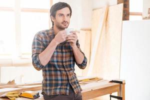 Cup of coffee for good work. Thoughtful young male carpenter holding coffee cup while leaning at the wooden table with diverse working tools laying on it photo