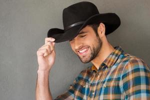 vaquero sonriente. apuesto joven ajustando su sombrero de vaquero y sonriendo mientras está de pie contra el fondo gris foto