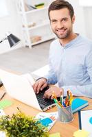 Love the job you do. Handsome young man in shirt working on laptop and smiling at camera while sitting at his working place photo