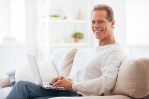 Enjoying leisure time at home. Side view of mature man working on laptop and smiling while sitting on the couch at home photo