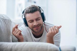 Enjoying his favorite music. Cheerful young man in headphones listening to the music and gesturing while lying on his couch at home photo