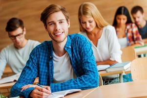 Showing a dedication to education. Handsome young man writing in note pad and looking at camera while sitting at front desk in the classroom photo